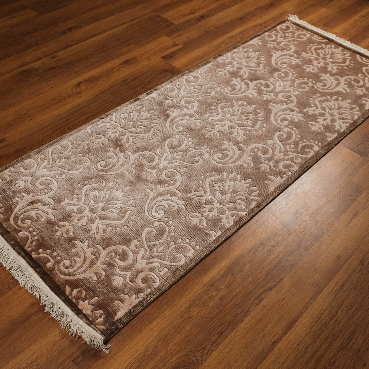 pudra damask desenli halı — koyu ahşap zeminde açılı duruş, ön köşede krem saçak